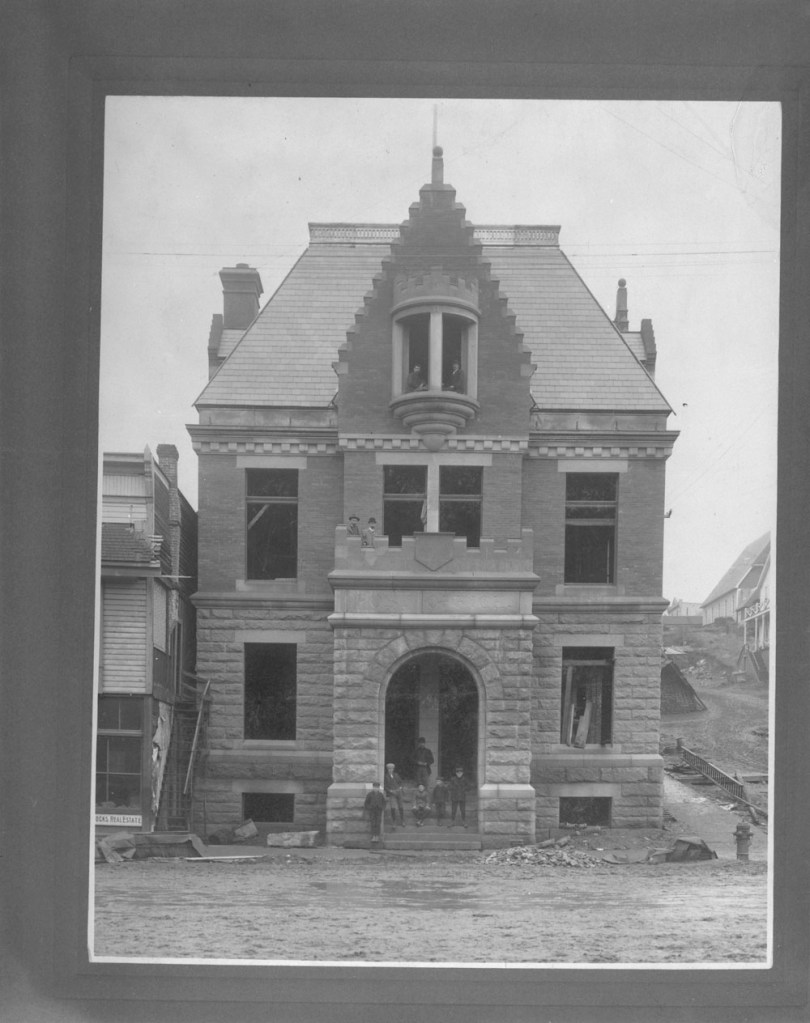 Black-and-white photograph of the exterior of the post office building. Small groups of men and children stand at the building’s unfinished entrance and one of its unfinished windows.