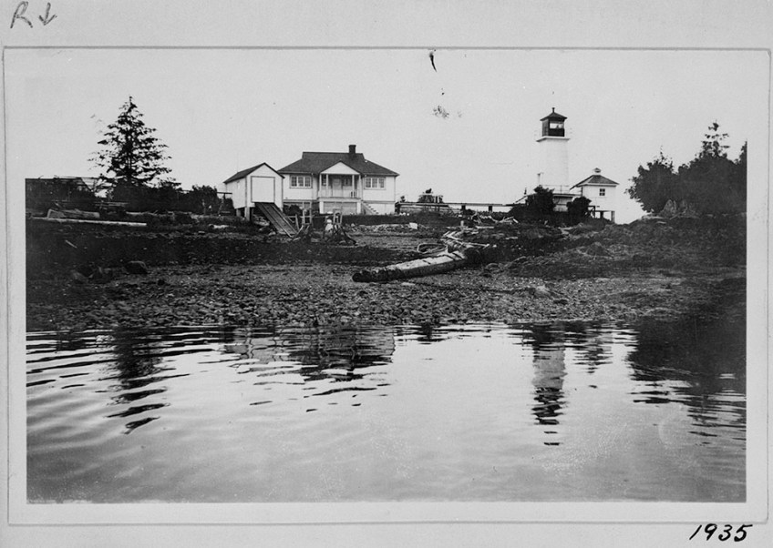 Black-and-white photograph of a lighthouse tower, a residence, a boathouse and other buildings, with the shoreline in the foreground.