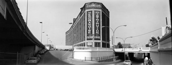 A black-and-white photograph of a tall factory building located at an intersection. There is a bridge on the right-hand side linking the building to a roadway. Cars are parked in a row on the left-hand side. 