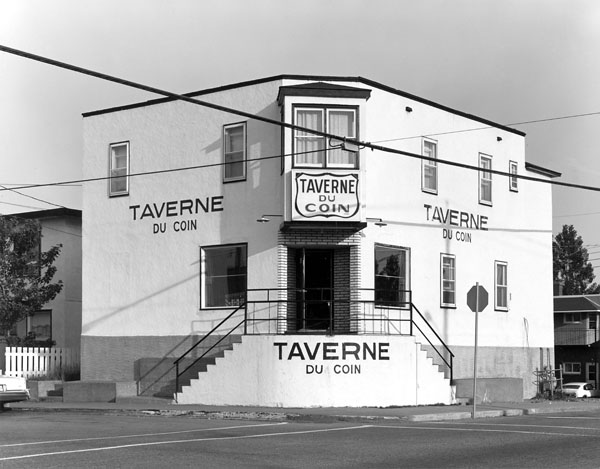 A black-and-white photograph of a white building with two sets of stairs leading to the entrance on a street corner. Written on the building in four places are the words “Taverne du Coin.” There is a stop sign near the building.