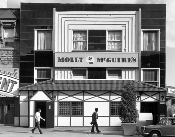 A black-and-white photograph of a building whose façade is white and dark. The words “Molly McGuire’s Pub” (with a small shamrock) are written. There are two men and a car in the foreground. 