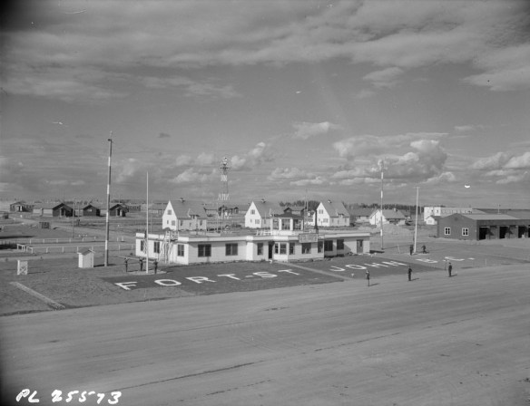 : Black-and-white photograph of the administration building at RCAF Fort St. John, B.C. Various individuals stand at attention as three men raise a flag. Several buildings and other structures are also in the background.