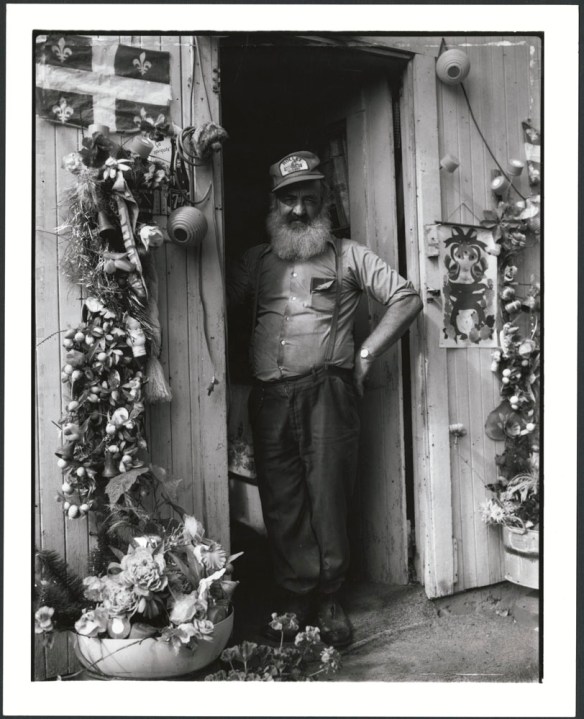 A man standing in a doorway, his left arm akimbo. He is wearing a ball cap and suspenders, and is smiling towards the camera. Flowers and a Quebec flag surround the door. 