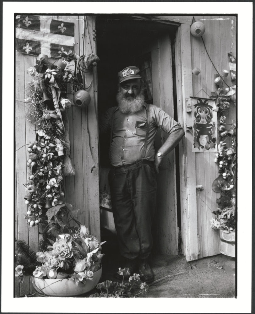 A man standing in a doorway, his left arm akimbo. He is wearing a ball cap and suspenders, and is smiling towards the camera. Flowers and a Quebec flag surround the door.