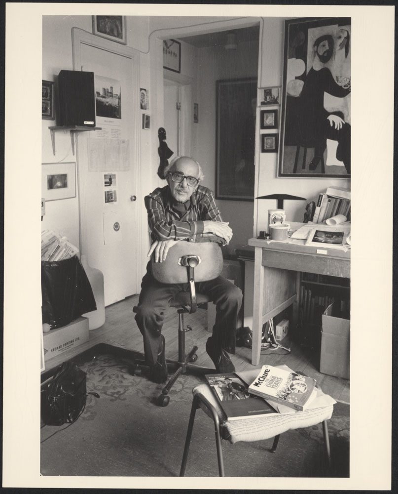 A black-and-white photograph of a man, in a striped buttoned-up shirt and dark trousers, looking towards the camera. He is sitting backwards on a rolling chair beside a desk, on which a lamp, papers and books can be seen.
