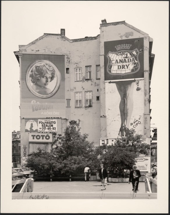 A black-and-white photograph of a tall old building with advertising featuring the Canada Dry logo, a woman with blond hair and a woman’s legs in high heel shoes. There are bushes in the front of the building, and people are moving about.
