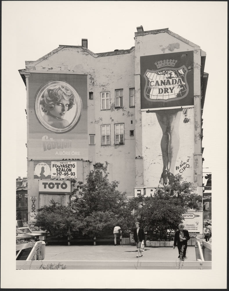 A black-and-white photograph of a tall old building with advertising featuring the Canada Dry logo, a woman with blond hair and a woman’s legs in high heel shoes. There are bushes in the front of the building, and people are moving about.