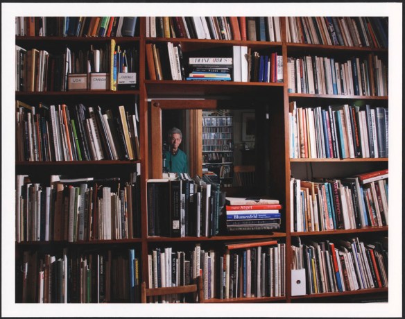 A colour photograph of Gabor Szilasi looking towards the camera, through a mirror, surrounded by shelves of books
