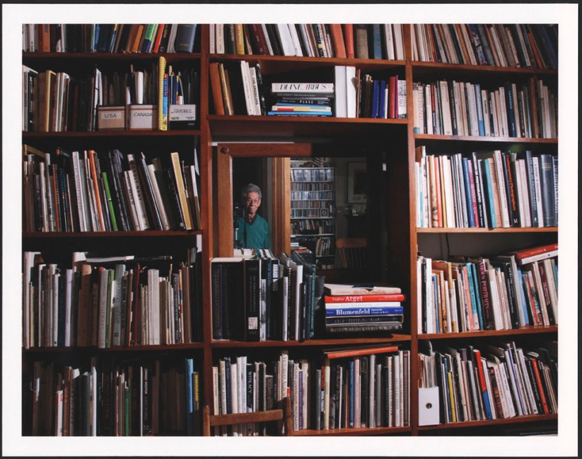 A colour photograph of Gabor Szilasi looking towards the camera, through a mirror, surrounded by shelves of books
