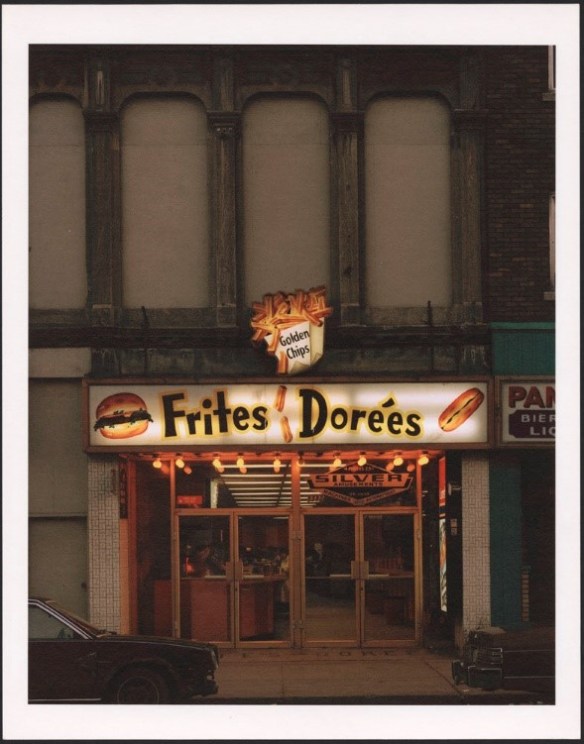 A colour photograph of a lit-up restaurant sign. The sign is on a brick building with large arches. The words “Frites Dorées” and images of a hamburger, a poutine and a hotdog can be seen on the sign. 