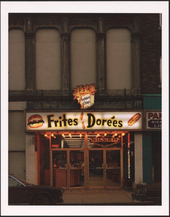 A colour photograph of a lit-up restaurant sign. The sign is on a brick building with large arches. The words “Frites Dorées” and images of a hamburger, a poutine and a hotdog can be seen on the sign.