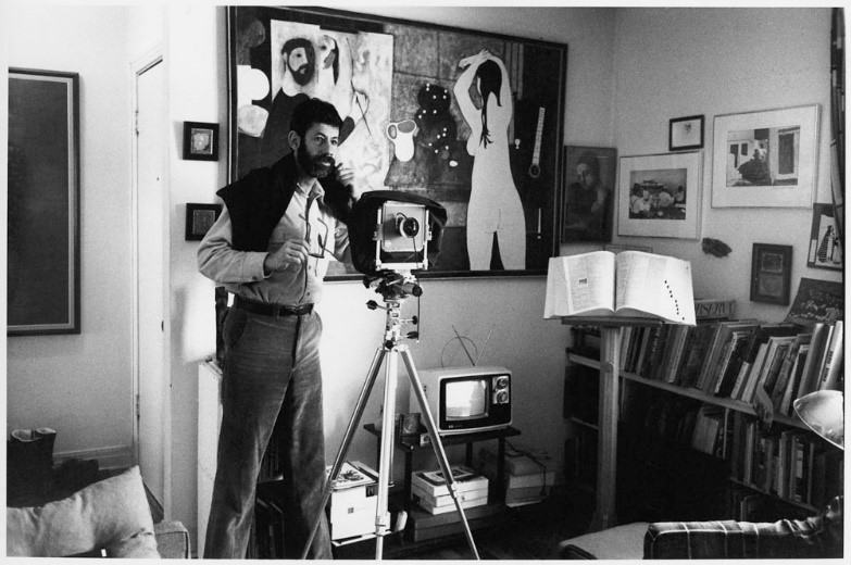 A black-and-white photograph of a man in a buttoned-up shirt, with a sweater over his shoulders. The man is standing behind a medium-format view camera.