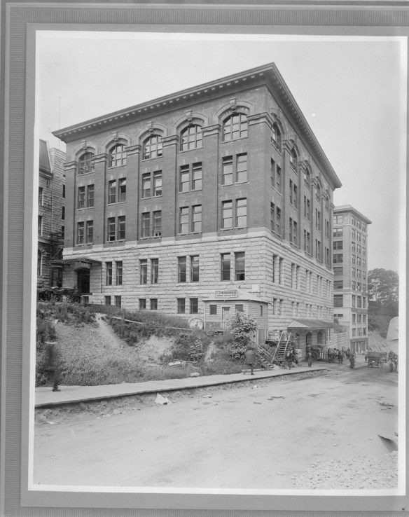 Black-and-white photograph of the exterior of the Customs Examining Warehouse in Vancouver, B.C. There is a wooden sidewalk in the foreground, and horse-drawn carts are delivering goods to the building entrance.