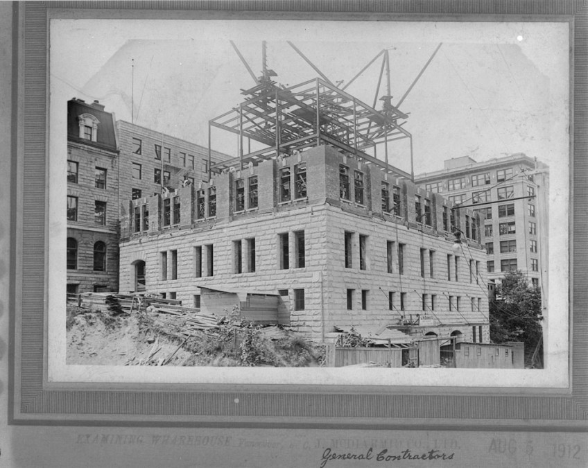 Black-and-white photograph showing the construction of the Customs Examining Warehouse in Vancouver. The lower section has a stone exterior, and the upper section has a steel structure and unfinished brickwork.