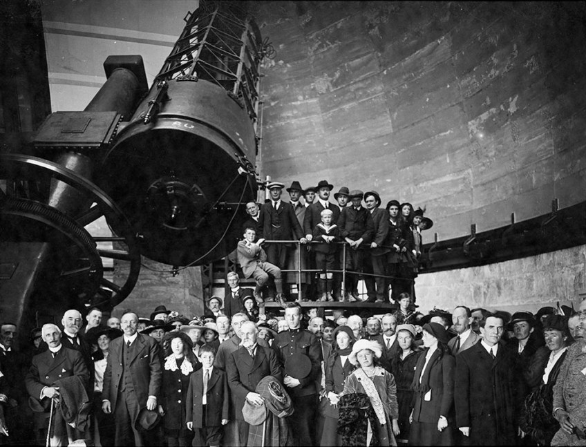 Black-and-white photograph of a large group of people inside the Dominion Astrophysical Observatory for its opening ceremony. The group is gathered in front of the telescope and on the viewing platform.
