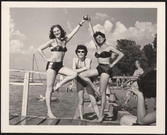 A black-and-white photograph of three women in bathing suits posing for the camera. They are standing on a dock by water.