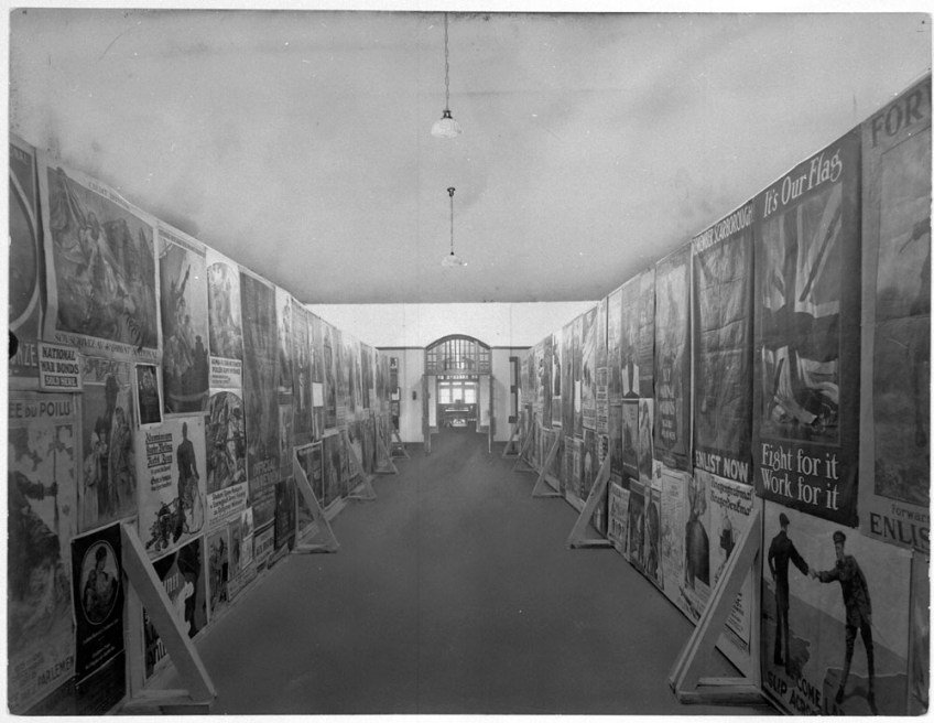 Black-and-white photograph of a long narrow room. There are two hanging lights in the middle of the room, and posters are plastered on each wall.