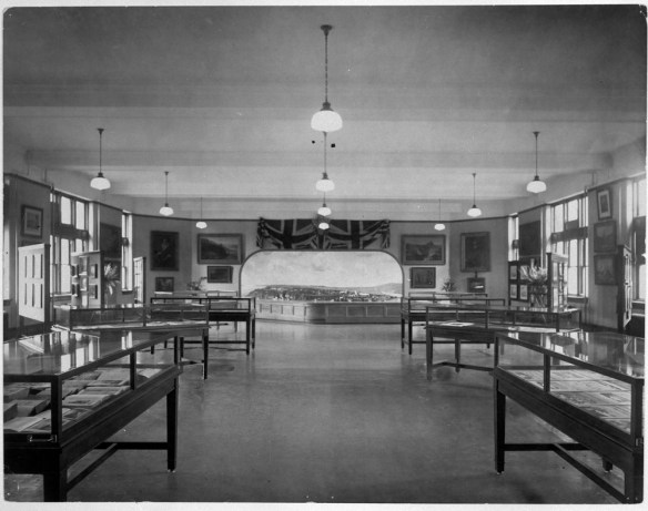 Black-and-white photograph of a large room with display tables running along the sides and lights hanging from the ceiling. In the background, there is a large model of a city. 