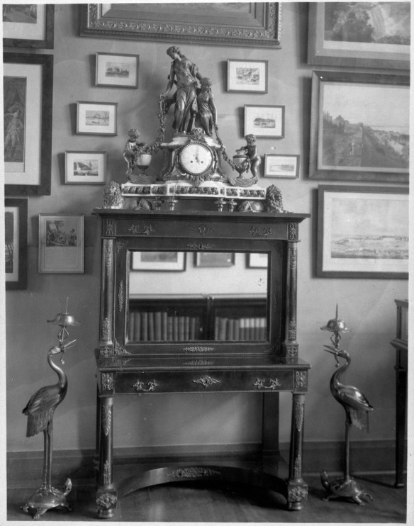 Black-and-white photograph of a wood cabinet with a mirror. On top of the cabinet is an elaborately decorated mantel clock. There are two candelabras in the shape of cranes standing on turtles’ backs on either side of the cabinet.