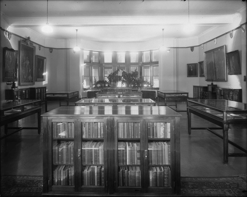Black-and-white photo of a room with book cases and display cabinets. In the background, there are windows and plants.