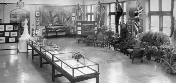 Black-and-white photograph of a large room with display cabinets, a statue, flags, plants, framed images, chairs and a throne.