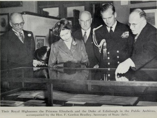 Black-and-white photograph of four men and one woman looking at items in a glass display cabinet. One man is pointing at a document in the cabinet.