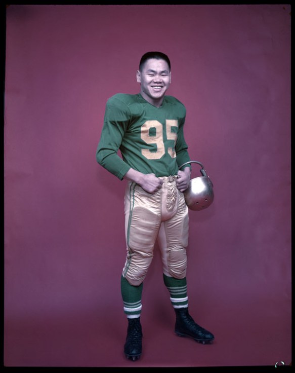 A colour studio photograph of a football player in uniform, holding his helmet in the crook of his left arm.