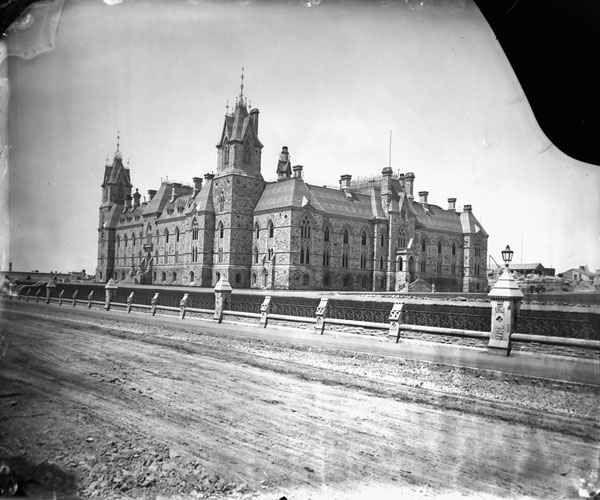 A large stone building with towers, behind a dirt road and a wrought-iron and stone fence.