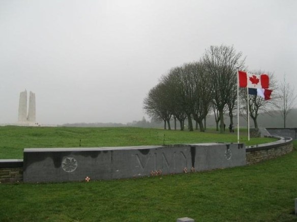 An expanse of green grass showing a white stone memorial in the distance, a grey stone sign with the engraved word VIMY and maple leaf symbols. The Canadian and French flags are on the right, against a foggy sky.