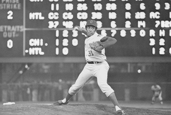A Black man in a white baseball uniform pitching a baseball, with a scoreboard behind him 