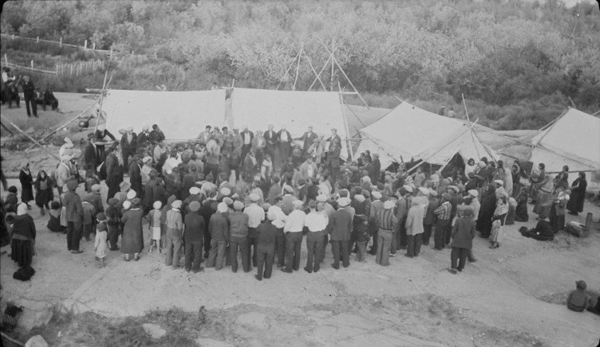 Black-and-white photograph from an elevated view showing a large group of people standing together in an elongated circular formation in the middle ground. Behind the group are four white canvas tents, and beyond the tents is a forest.