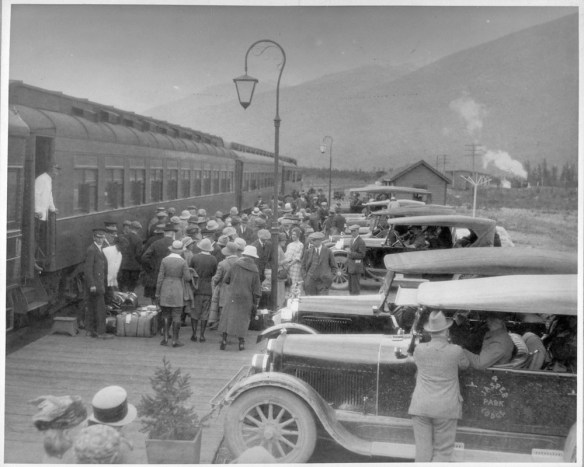 A crowd of people disembark from a train as railway employees and porters help them with their luggage.