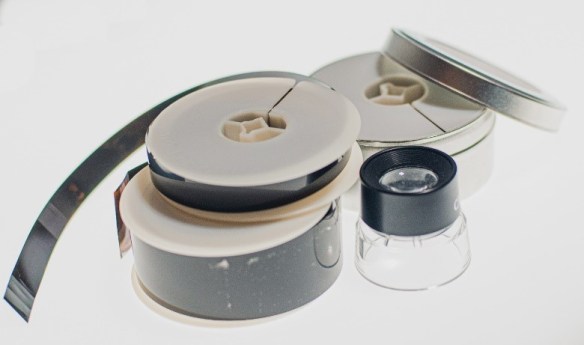 A photograph of a 16-mm microfilm reel stacked on top of a 35-mm microfilm reel, next to a loupe, in front of a 35-mm microfilm reel in its canister.