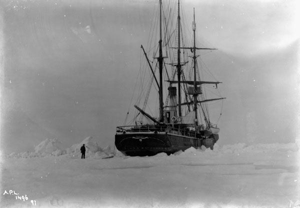 A black-and-white photograph of a square-rigged sailing ship with three masts surrounded by ice. A person stands on the ice to the left of the stern.