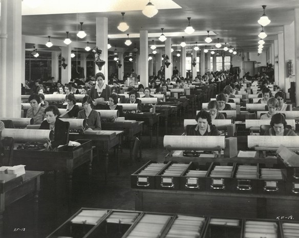 A black-and-white photograph of a large, high-ceilinged room in which approximately 80 employees (mostly women) sit at machines, punching in information recorded on a large roll in front of each machine. The employees sit in six columns, each of about a dozen rows, facing the photographer. The rows of employees are so numerous that they seem to extend into the distance. In the foreground, drawers of cards are lined up carefully on desks. In the middle ground, a woman stands observing a woman sitting at a punching machine. At the far right and in the background, additional personnel (mostly men) sit at additional rows of desks or are walking about.