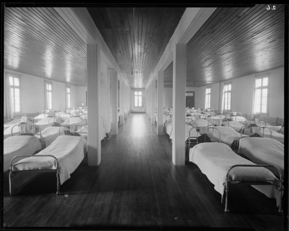 Black-and-white photograph of the interior of a large dormitory room, showing beds with white covers arranged sideways in two long rows on either side of an aisle.