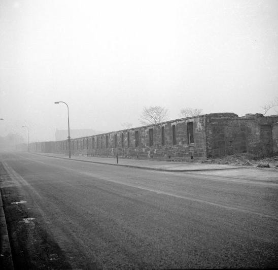 Black-and-white photograph of a paved street in front of a long unit of derelict row houses. The houses are built of stone and no longer have roofs.