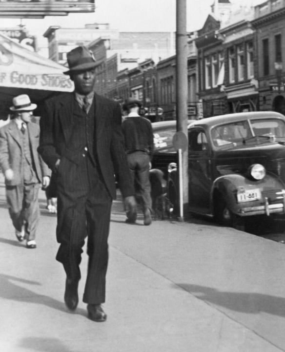 A man in a suit and hat walking down a sidewalk lined with cars and buildings.