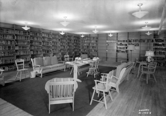 Chairs circling a table in the middle of a room surrounded by shelves with books.