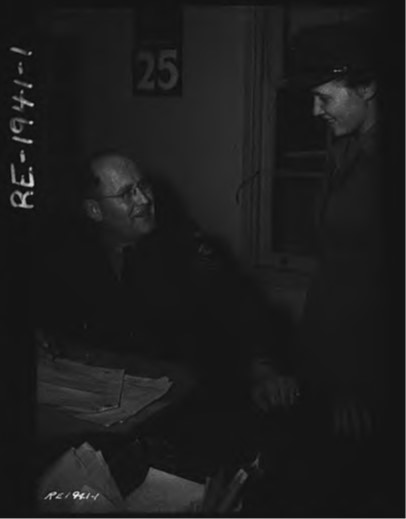 A seated uniformed man wearing glasses (left) looks towards a standing uniformed woman (right).