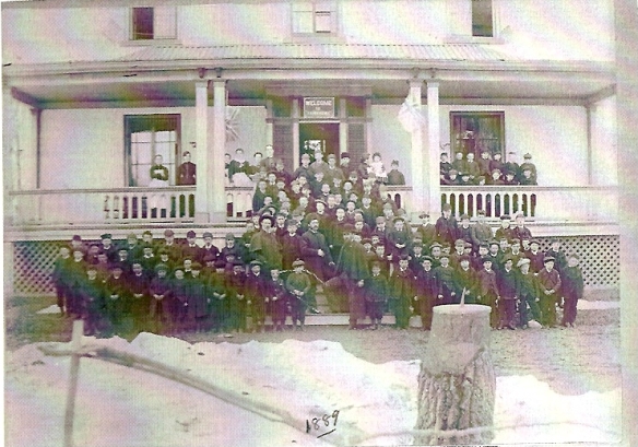 Black and white photograph of a large group of boys, several men and women standing on the ground, front steps, and verandah of a white stucco building.