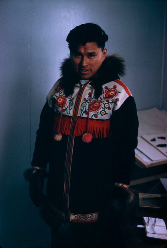 A colour portrait photograph of Abe Okpik standing indoors wearing a black Inuit parka with yoke and red, yellow and green floral embroidery on a white yoke with red fringe. He has black fur mitts on.