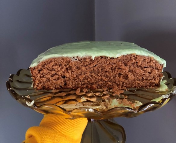 A cross section shot of a brown cake topped with green frosting. The cake sits on a glass platter.