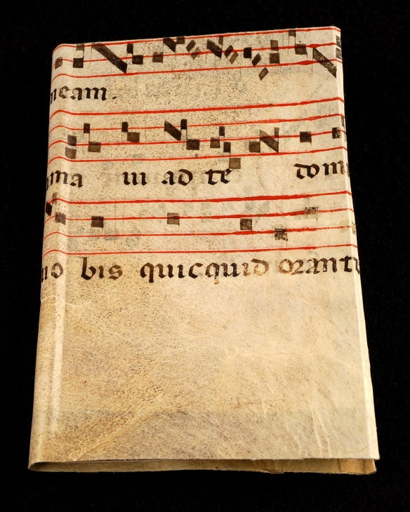 A photograph showing a small book bound in a piece of vellum with musical notations handwritten on it in red and black ink in a calligraphic style. The vellum appears to date from the sixteenth or seventeenth century.