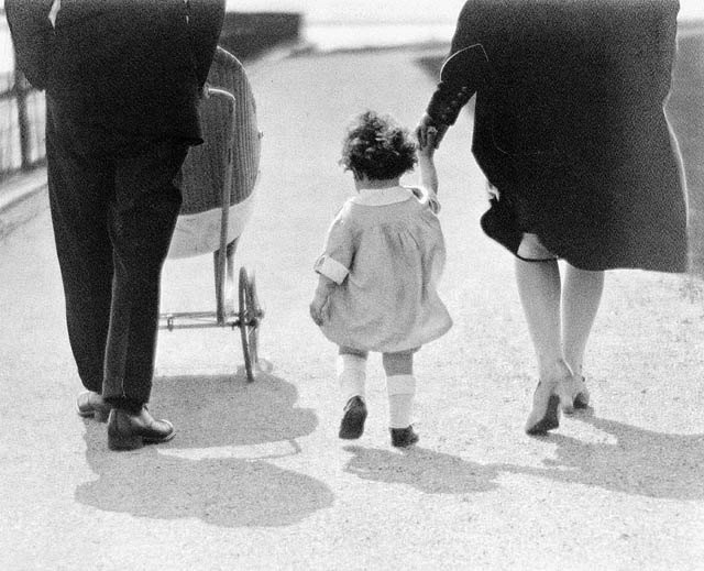 Black-and-white photograph featuring three people walking from behind. A man on the left pushes a stroller, while a small child in the centre holds the hand of a woman on the right.