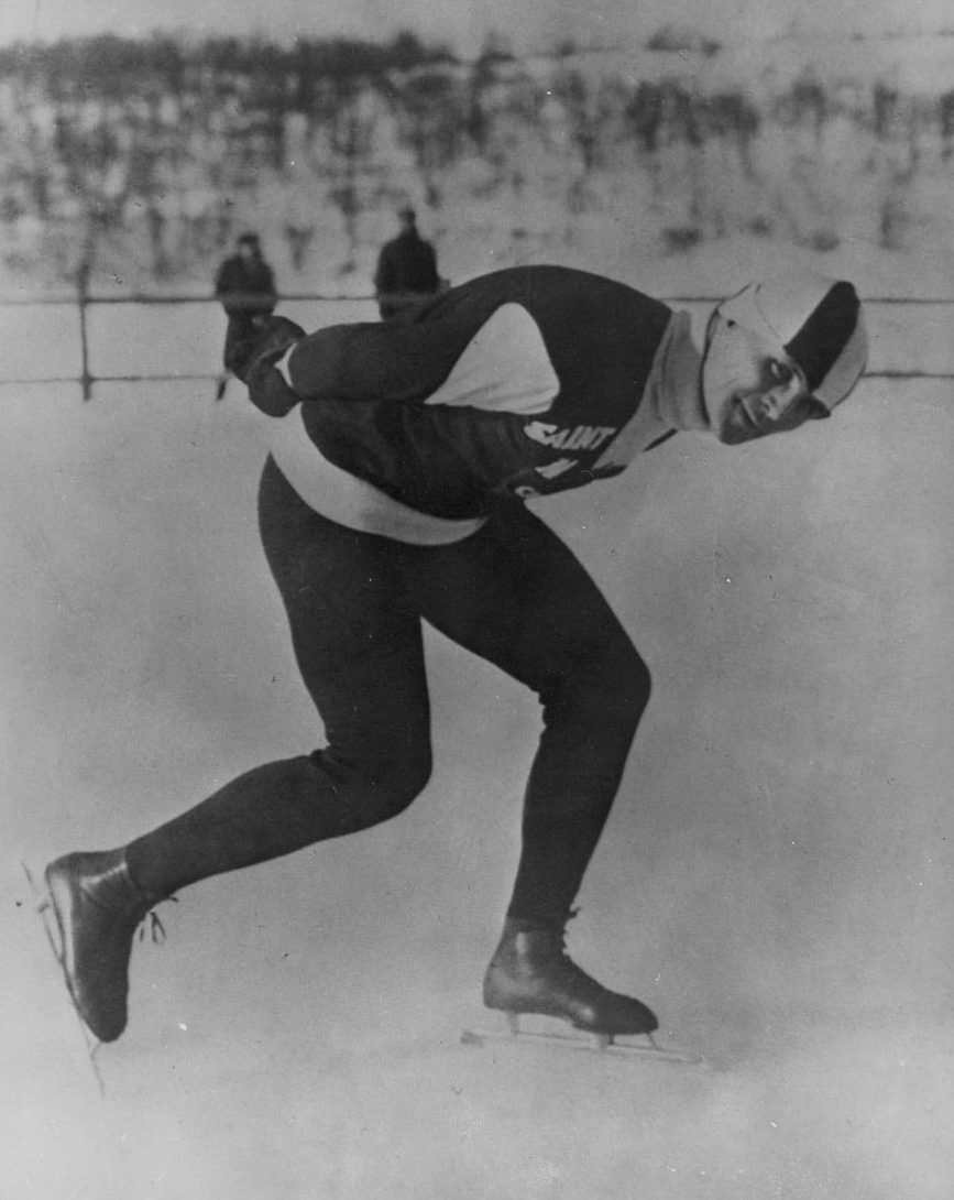 A black and white photograph of a man speed skating and looking directly at the camera.