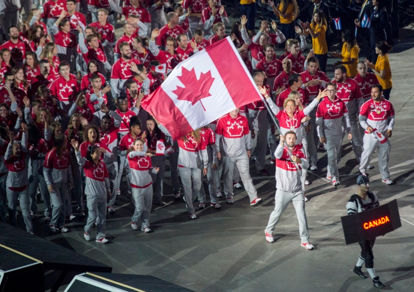 Photograph of numerous athletes marching behind a man carrying a large Canadian flag.