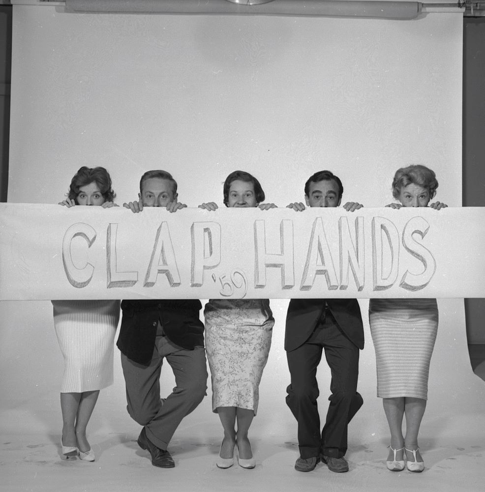 Image of five people taking a Kilroy Was Here pose gripping a banner with the words CLAP HANDS in front of them.